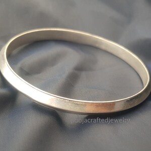 Puede incluir: Un brazalete plateado con un diseño liso y redondeado. El brazalete circular parece estar hecho de metal. La joya se exhibe sobre un fondo de tela azul oscuro. El brazalete es un accesorio sencillo y elegante.