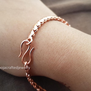 May include: A copper-colored bracelet with a textured, scale-like design. The bracelet is secured with a hook clasp. The jewelry is shown on a person's wrist.