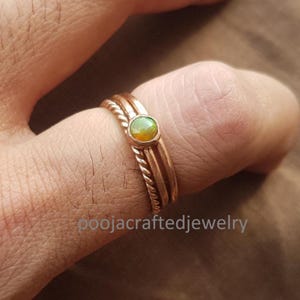 Pode incluir: Um conjunto de três anéis de cor cobre. Um anel apresenta uma pequena gema de opala redonda. Os outros dois anéis têm um design de corda torcida. Os anéis estão em um dedo. O texto "poojacraftedjewelry" é visível.