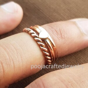 May include: Three copper-coloured rings are stacked on a finger. The rings include a twisted band, a smooth band, and a band with a small heart-shaped charm. The rings are displayed on a brown background. The text "poojacraftedjewelry" is visible.