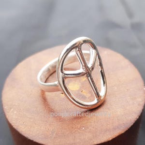 May include: A silver-colored ring featuring a peace symbol design. The ring has a polished finish and is set on a wooden surface. The ring's design is a prominent, openwork peace symbol.