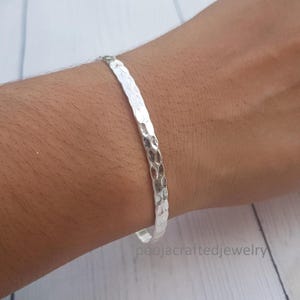 May include: A silver-colored bangle bracelet featuring a hammered, textured surface. The metal band has a reflective finish and a consistent width throughout its circular design. The text poojacraftedjewelry is visible in the lower right corner.