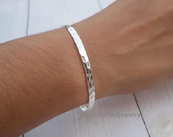 May include: A silver-colored bangle bracelet featuring a hammered, textured surface. The metal band has a reflective finish and a consistent width throughout its circular design. The text poojacraftedjewelry is visible in the lower right corner.