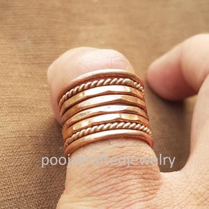 May include: Copper rings stacked on a finger. The rings have a hammered texture and a twisted rope design. The rings are a warm copper color. The text "poojacraftedjewelry" is visible.