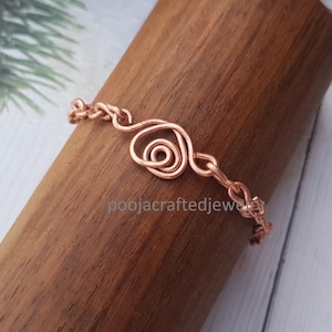 May include: A copper-colored bracelet with a spiral design in the center. The bracelet is displayed on a wooden stand. The text "poojacraftedjewelry" is visible on the stand.