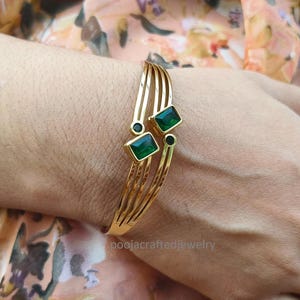 Könnte beinhalten: Ein goldfarbenes Armband mit vier parallelen Bändern, die sich in zwei Abschnitte teilen. Das Design ist mit zwei rechteckigen smaragdgrünen Steinen und zwei kleinen runden schwarzen Steinen akzentuiert, was eine symmetrische, offene geometrische Optik erzeugt.