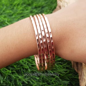 Pode incluir: Um conjunto de seis pulseiras de cobre martelado. As pulseiras têm uma superfície texturizada e uma cor quente de ouro rosa. São exibidas em um braço contra um fundo de grama verde. O nome da loja, poojacraftedjewelry, é visível.