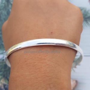 May include: A silver bangle bracelet. The smooth, rounded design of the bracelet reflects light, creating a bright, shiny appearance. The jewellery is a simple, elegant accessory.