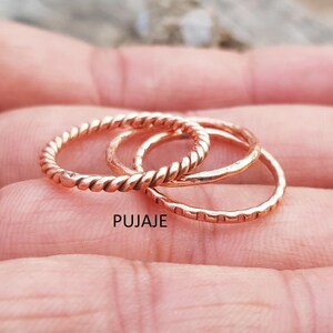 Op de afbeelding: Drie koperkleurige ringen. Eén ring heeft een gedraaid touwontwerp, een andere is een eenvoudige band en de derde heeft een reeks kleine rechthoekige inkepingen. Het woord "PUJAJE" staat in de afbeelding.