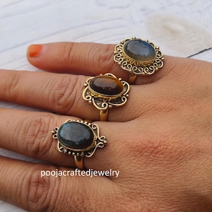 Puede incluir: Tres anillos de tono dorado con piedras preciosas ovaladas en cabujón en tonos gris, marrón y azul oscuro. Cada anillo tiene un engaste de metal decorativo intrincado con detalles de filigrana alrededor de la piedra central. Texto poojacraftedjewelry.