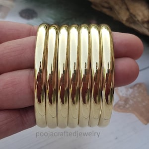 May include: A set of seven gold-colored bangles. The bangles are smooth, with a rounded shape and a polished finish. The jewelry is held in a hand, with a blurred background.