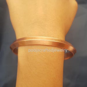 May include: A copper-coloured bangle bracelet with a smooth, polished finish. The bracelet is a solid band with a slightly angular design. The text "poojacraftedjewelry" is visible on the wrist. This piece of jewellery is a simple, elegant accessory.