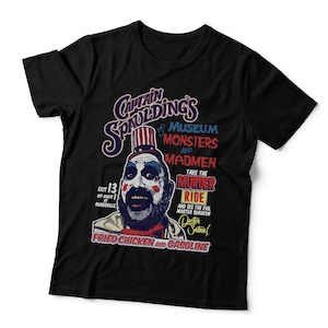 May include: A black short-sleeved t-shirt featuring a graphic of Captain Spaulding wearing a top hat. The text reads: Captain Spaulding's Museum of Monsters and Madmen, Take the Murder Ride, Exit 13, Fried Chicken and Gasoline, and Doctor Satan.