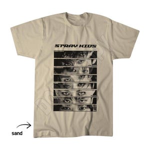 May include: A sand-coloured short-sleeve t-shirt featuring the text STRAY KIDS at the top. Below the text are six horizontal rectangular panels, each displaying a close-up of a pair of eyes, with individual names printed next to each set of eyes.