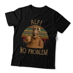 May include: A black short-sleeved t-shirt featuring a graphic of the character Alf giving a thumbs up. The design includes a sunset-coloured striped circle background, a forest silhouette, and the text "ALF!" above and "NO PROBLEM" below the character.