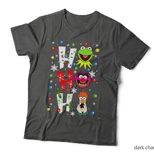 May include: A dark charcoal grey short-sleeve t-shirt featuring the text HO HO HO with the letters integrated into a Muppets graphic. The design includes Kermit the Frog, Animal, and Beaker, surrounded by colourful Christmas lights and snowflakes.