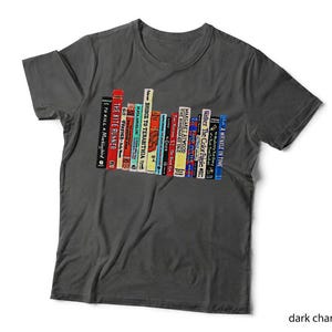 May include: A dark charcoal grey short-sleeve t-shirt featuring a graphic print of a row of colourful book spines. The books display various titles including To Kill a Mockingbird, The Kite Runner, Bridge to Terabithia, and A Wrinkle in Time.