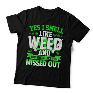May include: A black short-sleeved t-shirt featuring a graphic print in green and white. The text reads YES I SMELL LIKE WEED AND YOU SMELL LIKE YOU MISSED OUT, accompanied by illustrations of cannabis leaves and two rolled smoking items.