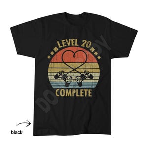 May include: A black short-sleeved t-shirt featuring a graphic print of two game controllers with cords forming a heart shape. The design includes a sunset-coloured circle background with horizontal stripes and the text LEVEL 20 COMPLETE.