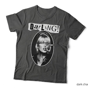 May include: A dark charcoal grey t-shirt featuring a black and white oval graphic of a person with blonde hair and a cigarette. Above the oval is the text "Darling!" in a torn paper style, and across the eyes is the text "Absolutely Fabulous".