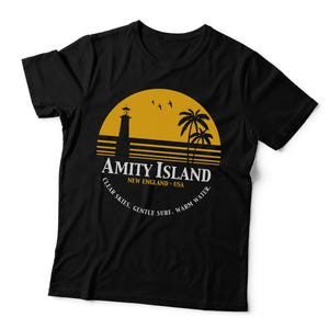 May include: A black short-sleeved t-shirt featuring a yellow circular graphic of a lighthouse, palm trees, and birds. The text reads AMITY ISLAND NEW ENGLAND - USA and CLEAR SKIES. GENTLE SURF. WARM WATER. The garment is a solid black colour throughout.