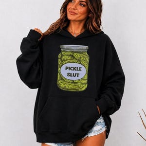 May include: A black hooded sweatshirt featuring a graphic print of a glass jar filled with green pickles. The jar has a light blue oval label on the front with the black text PICKLE SLUT printed in the centre. The hoodie has long sleeves and a front pocket.