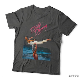 May include: A dark charcoal grey short-sleeve t-shirt featuring a graphic print of a person lifting another person above their head in water. The text Dirty Dancing is printed in a stylised pink script font above the central photographic image.