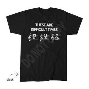 May include: A black short-sleeved t-shirt featuring white text that reads "THESE ARE DIFFICULT TIMES" above three musical staves, each displaying a treble clef and the time signatures 13/8, 11/4, and 7/16 in a white font on the chest area.