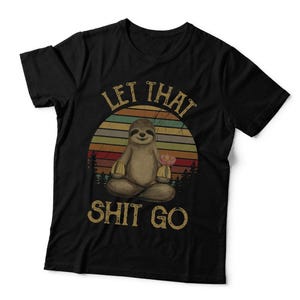 May include: A black short-sleeved t-shirt featuring a graphic of a sloth sitting in a lotus position. The design includes a sunset background with horizontal stripes in muted colours, a forest silhouette, and the text "LET THAT SHIT GO" in a distressed, vintage-style font.