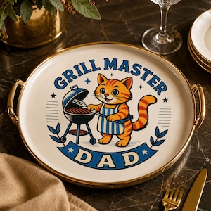 Può includere: Un vassoio rotondo con manici dorati e superficie bianca. Il vassoio presenta un gatto dei cartoni animati che griglia, con le parole "GRILL MASTER DAD" in blu. Il gatto è arancione e indossa un grembiule a righe blu e bianche.