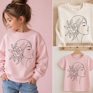 May include: A light pink sweatshirt, a cream-colored sweatshirt, and a light pink t-shirt. Each garment features a black line drawing of a woman's profile with a floral hairpiece and earrings. The t-shirt is on a wooden hanger.