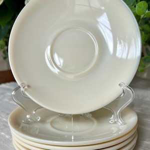 May include: A stack of five round, off-white milk glass saucers with a smooth, glossy finish. Each saucer features a slightly raised circular center indentation designed to hold a cup securely. The plates have a simple, clean edge design.