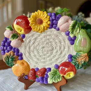 May include: An oval ceramic serving platter featuring a central cream-colored basket-weave pattern. The rim is decorated with three-dimensional sculpted fruits and a sunflower, including red apples, purple grapes, a green pear, and strawberries.