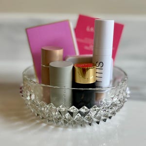 May include: A clear, faceted glass bowl filled with beauty products. The bowl contains several cylindrical containers with gold, silver, and black caps, along with pink cards and a white tube with black text. The products are arranged on a white surface.