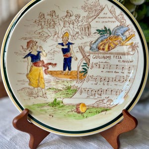 May include: A round ceramic plate with a cream background and a dark green rim. The surface features a colorful illustration of figures in a boat, a tent labeled Au Rendez-vous des Canotiers, and musical sheet music for Guillaume Tell by G. Rossini.