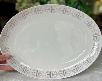 May include: An oval-shaped white ceramic serving platter featuring a repeating geometric pattern in a muted taupe color along the rim. The design consists of interconnected circular motifs that create a continuous border around the smooth center.