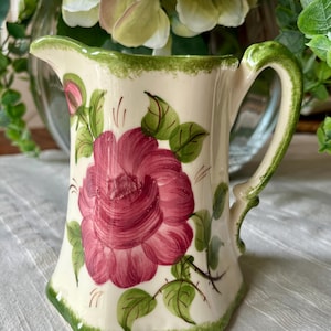 May include: A cream-colored ceramic pitcher featuring a hand-painted large magenta rose with green leaves and stems. The rim and base are accented with a textured green sponge-painted border. The pitcher has a curved handle and a small pouring spout.
