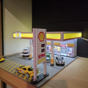 May include: A miniature Shell gas station model with a yellow and white canopy, a price sign, and several toy cars. The sign displays fuel prices. A yellow sports car with the number 95 is parked on the road.