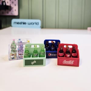 May include: Miniature soda bottles in plastic carriers. The carriers are red, blue, and green, with the Coca-Cola, Pepsi, and Sprite logos. One carrier has Hello Kitty bottles. The background includes a sign that says "meshie world".