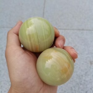 Peut inclure: Deux pierres sphériques polies avec une finition lisse et brillante. Les pierres sont de couleur vert clair et présentent des bandes translucides subtiles avec de légères stries de jaune pâle et de blanc sur la surface minérale.