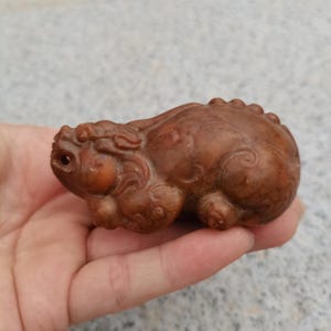Puede incluir: Pequeña figura tallada en marrón que representa una criatura mítica, posiblemente un perro Foo o una figura de guardián similar. La escultura tiene detalles, incluyendo una cara, un cuerpo y elementos decorativos definidos. La figura se sostiene en una mano.