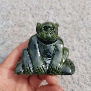 Puede incluir: Una figura de mono de jade tallado en posición sentada. El mono es de color verde oscuro con detalles tallados visibles. La figura se sostiene en una mano sobre un fondo gris claro.