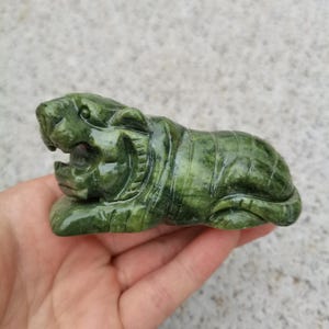 Könnte beinhalten: Eine geschnitzte grüne Jade-Tigerfigur, die mit offenem Maul liegt. Der Tiger ist mit geschnitzten Streifen und Gesichtszügen detailliert. Die Figur wird in einer Hand gehalten, um die Größe und die Details der Skulptur zu zeigen.
