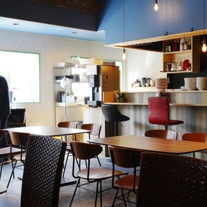 Może przedstawiać: Wnętrze restauracji z drewnianymi stołami i krzesłami. Osoba stoi blisko okna. Bar ma czerwone krzesło i czarne stołki. Nad głową wiszą żarówki.