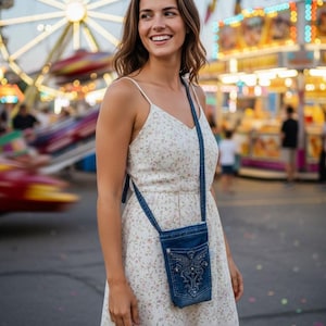 May include: A woman wearing a white floral print dress with spaghetti straps and a denim crossbody bag with decorative stitching. The background features a lit-up Ferris wheel and carnival lights.