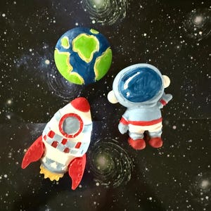 May include: A colorful space-themed collection featuring a hand-painted Earth, an astronaut, and a rocket ship. The Earth is blue and green, the astronaut is in a blue suit with a red stripe, and the rocket is white with red fins. The background is a starry night.