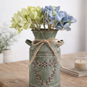 Peut inclure: Vase à fleurs rustique vintage avec des fleurs vertes et bleues. Le vase en métal vert olive a un motif de cœur et un nœud en ficelle. Le texte "Vintage Rustic Flower Vase" est en haut.