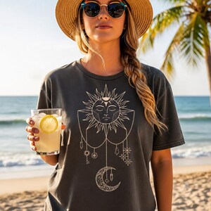 Pode incluir: Uma t-shirt de manga curta cinzento-carvão com um gráfico grande e detalhado de linhas brancas no peito, representando um sol com rosto, uma lua crescente, cristais pendentes, estrelas e uma chave.