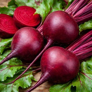 May include: Close-up of fresh beets with vibrant red-purple bulbs and green leaves. One beet is sliced, revealing a deep red interior. The beets are arranged on a wooden surface, showcasing their natural colors and textures.