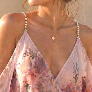 May include: A light pink floral print dress with a V-neckline and off-the-shoulder sleeves. The dress has pearl-like straps and a flowing design. A gold necklace with a small pendant is visible. The dress is made of a sheer fabric.
